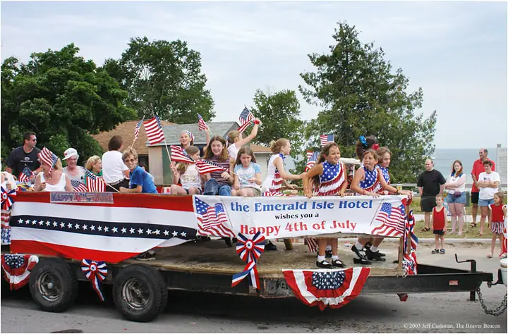 A patriotic parade float carries people and American flags past spectators on a summer street.