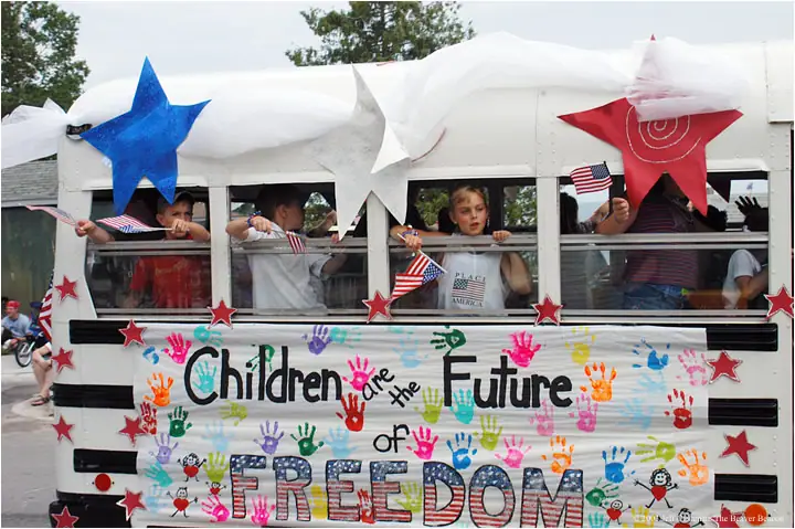 Children wave American flags from a decorated school-bus parade float covered with stars, handprints, and a freedom message.