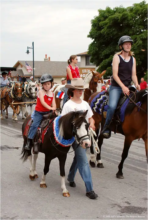 A mounted parade passes along a street, with children and adults riding decorated horses and ponies.