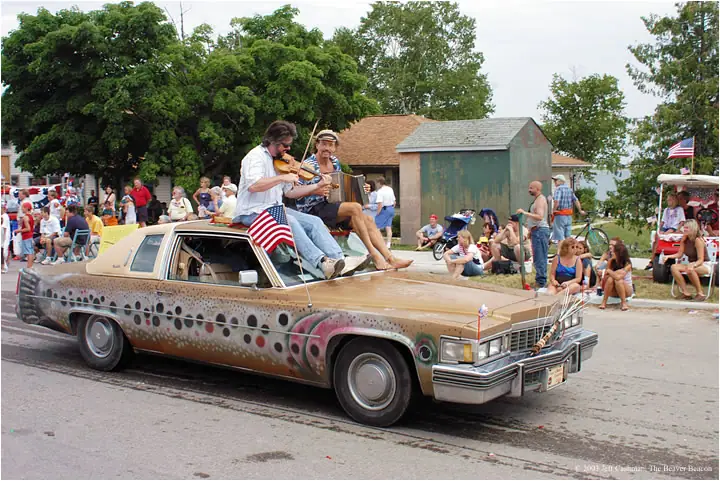 A decorated sedan carries two musicians past spectators gathered along a street during a parade.