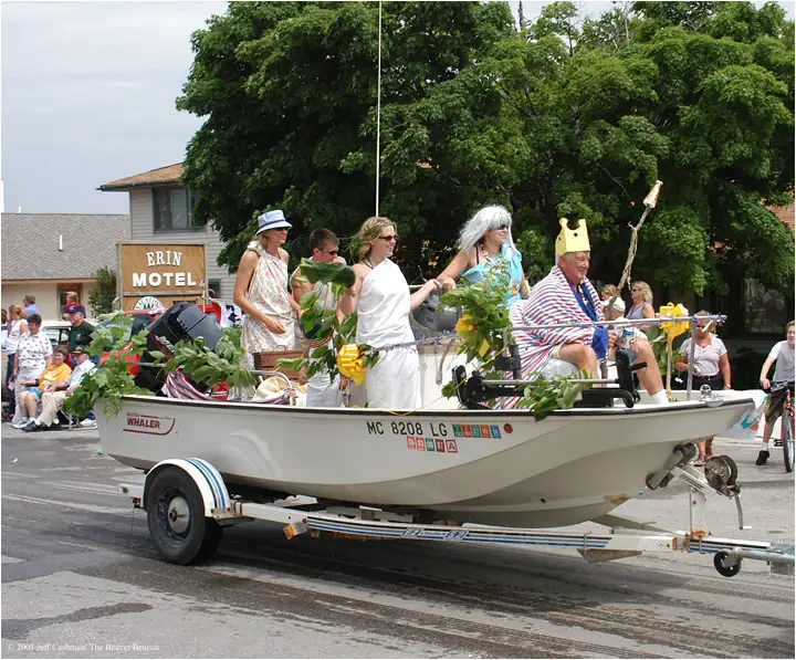 Costumed adults ride in a decorated white motorboat on a trailer during a street parade.