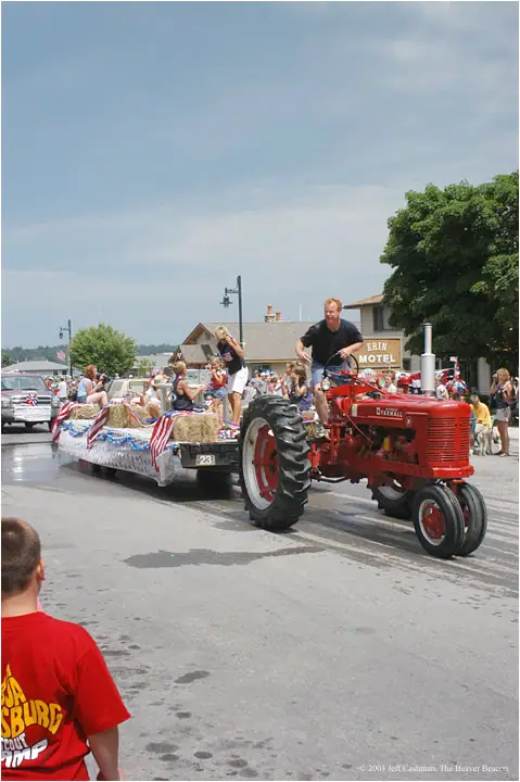 A red tractor pulls a flag-decorated hay wagon through a street parade as spectators watch.