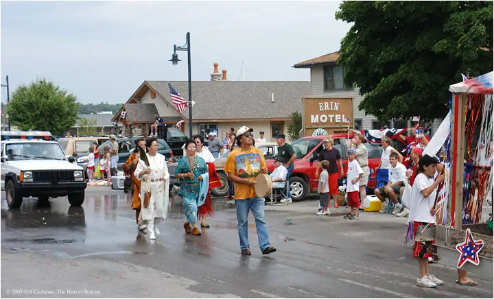 A rainy parade passes a motel, with a drummer and costumed participants walking among parked vehicles.