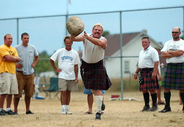 A kilted athlete throws a heavy stone while several people watch behind him.