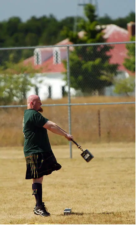 A kilted athlete swings a hammer across a dry grass field beside a chain-link fence.