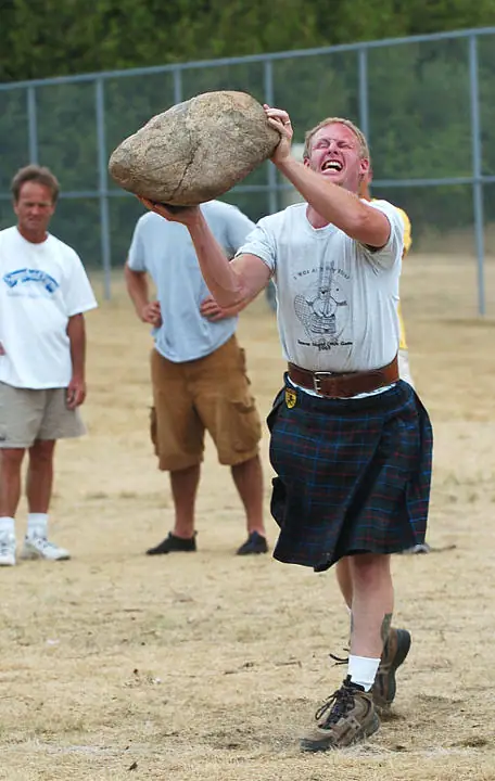 A man in a tartan kilt lifts a large stone while two men watch from a sandy field.