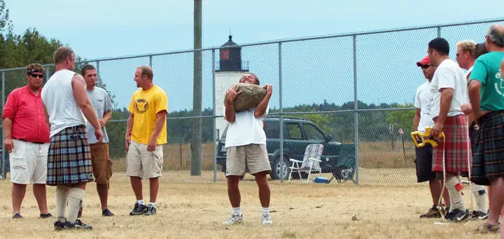 Men in kilts and casual clothing gather as one competitor lifts a large stone in a fenced field.