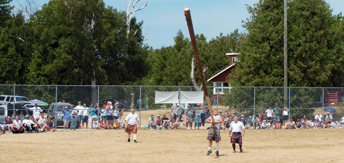 A kilted athlete raises a long wooden pole before spectators at an outdoor field event.
