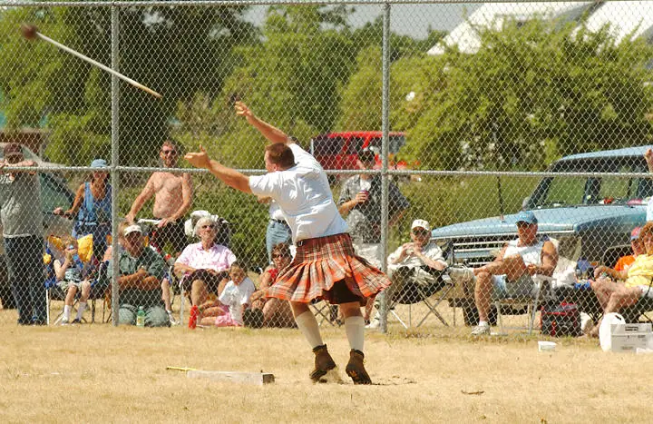 An athlete in a kilt throws a hammer before seated spectators behind a chain-link fence.