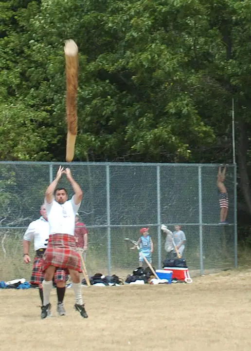 A kilted athlete throws a large wooden pole while spectators watch behind a chain-link fence.