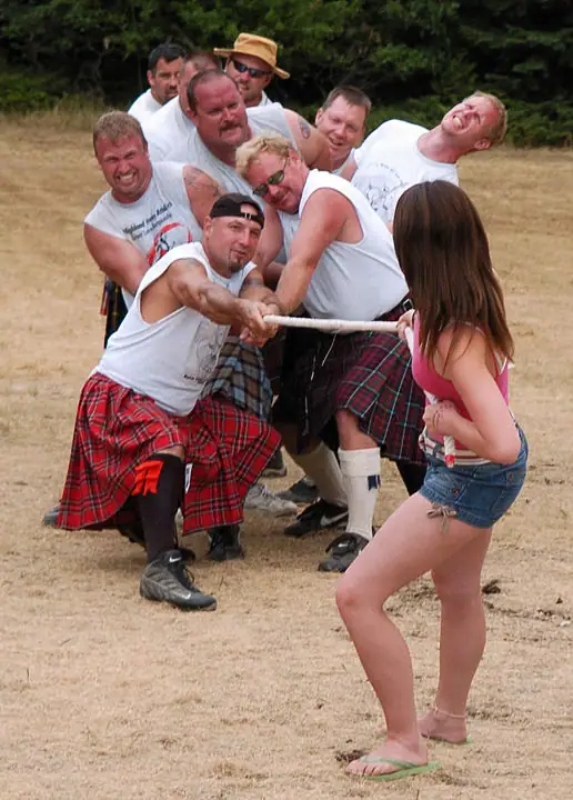 Men in tartan kilts pull a rope in a tug-of-war while a woman watches in the foreground.