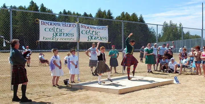 Outdoor games event with kilted performers, a bagpiper, children, spectators, and banners behind a fenced field.