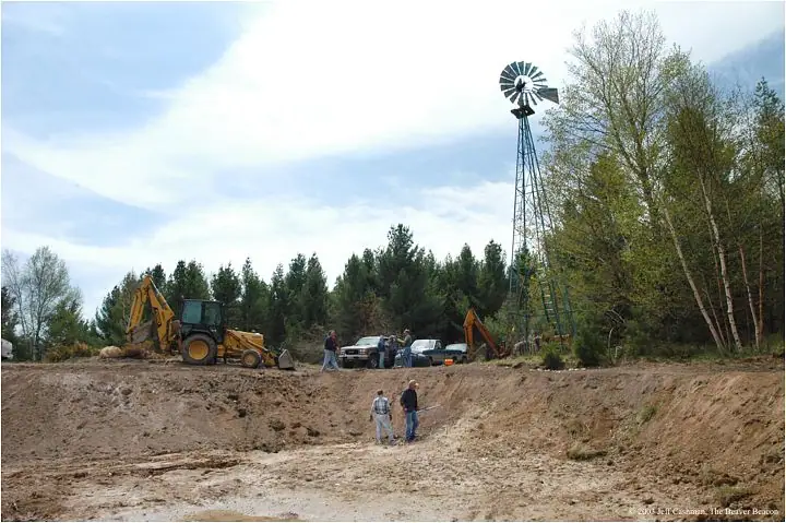 People stand in a large excavation beside construction equipment, pickup trucks, and a tall windmill.
