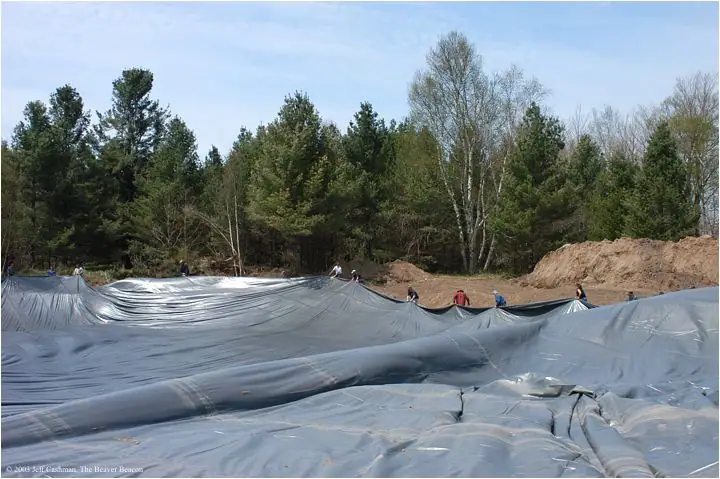 Workers spread a large black plastic liner across an excavated area beside a forest.