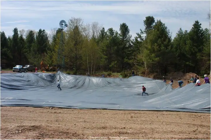 People spread a gray liner across an excavated pond beside a forest and windmill.