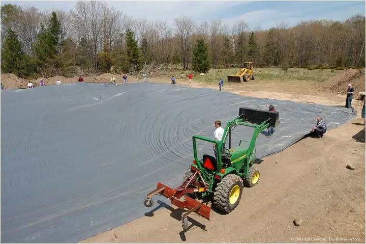 Workers spread a large dark liner over a pond basin beside a green tractor and yellow backhoe.