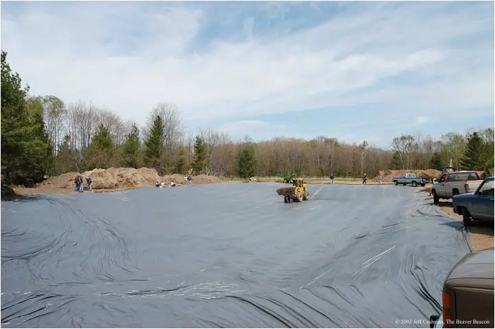 Lined pond construction site with a skid-steer loader, workers, pickup trucks, and a wooded background.