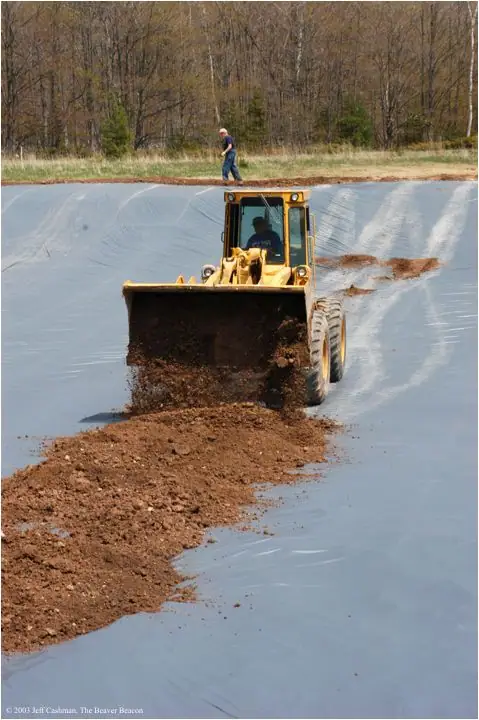 A yellow front-end loader spreads soil across a plastic-lined excavation while a person walks behind it.