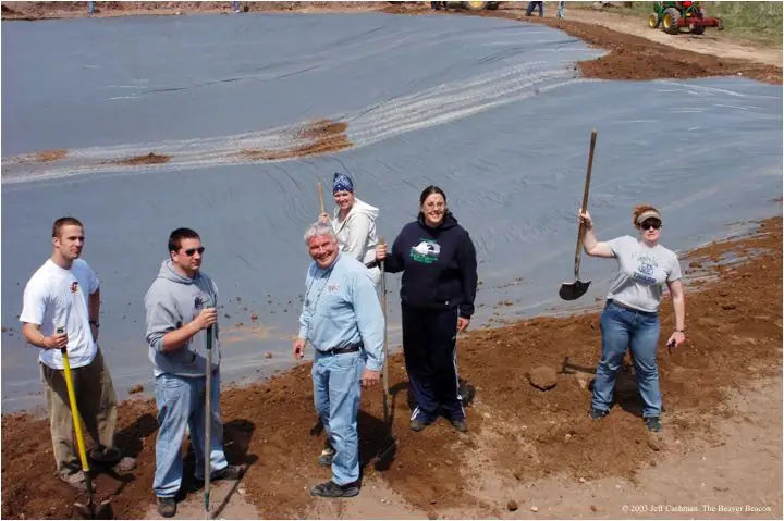 Six people with shovels stand beside a large pond lined with gray sheeting.