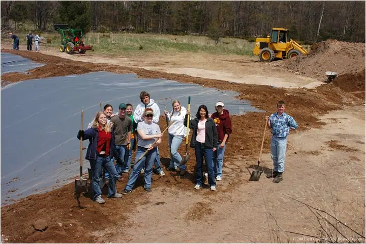 People pose with shovels beside a dark-lined pond under construction, with heavy equipment and woods behind them.