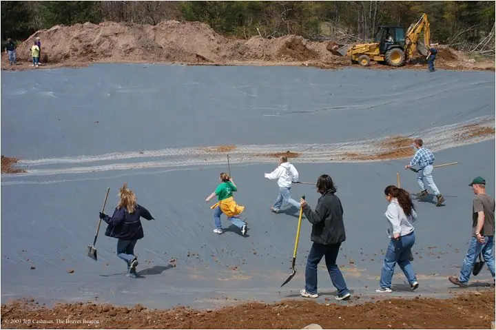 People spread soil across a large gray pond liner while a backhoe sits beside piles of earth.