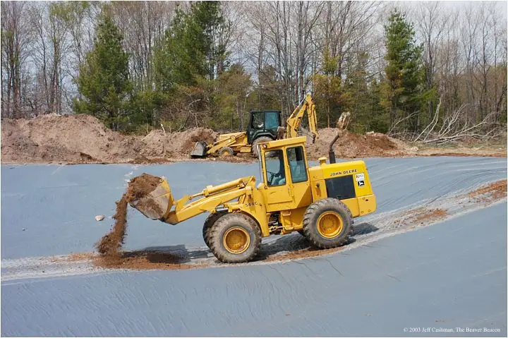 Two yellow earthmoving machines work over a broad blue-lined excavation beside a forest.