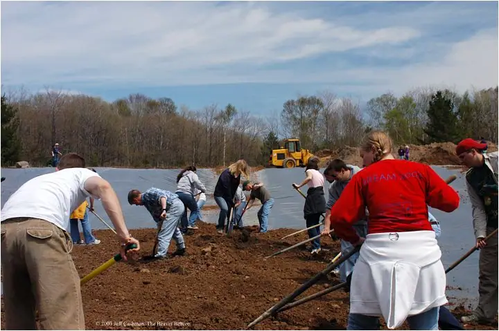 People use shovels and rakes to spread soil over a lined pond beside a front-end loader.