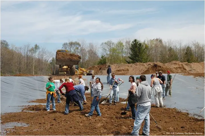 People spread soil over a large lined pond basin while dump trucks and construction equipment stand behind them.