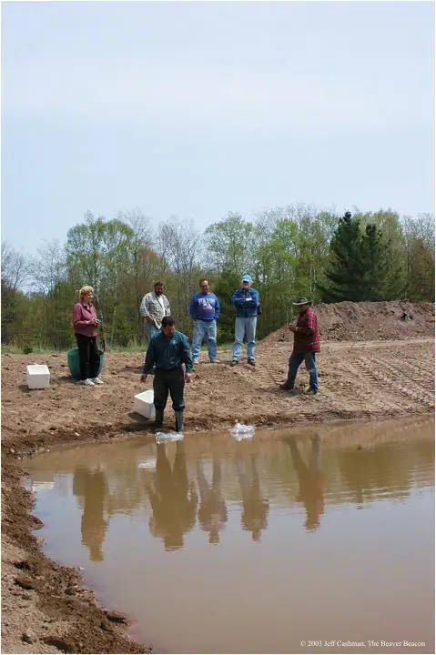 Six people work beside a muddy pond, with containers, disturbed soil, tire tracks, and a forest behind them.