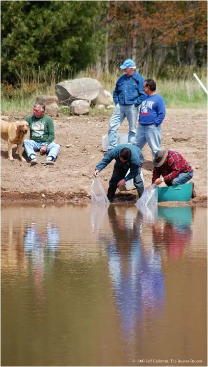 Five people and a dog gather beside a pond while two people collect water in clear bags.