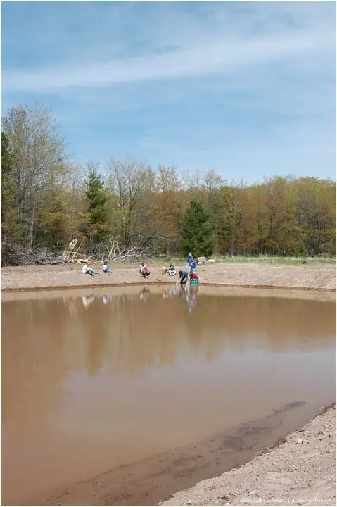 People gather beside a muddy, partially excavated pond bordered by forest.