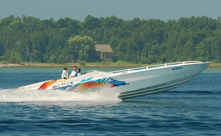 A white speedboat carrying three people speeds past a forested shoreline, throwing up a broad wake.