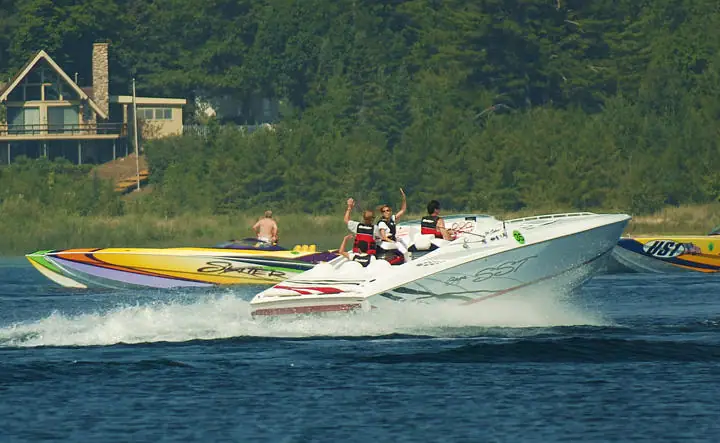 High-performance boats speed across the water, with passengers aboard the lead boat and a wooded shoreline behind.