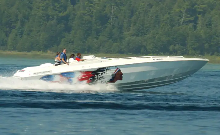 A white speedboat carrying four people speeds across the water, leaving a broad wake before a wooded shoreline.