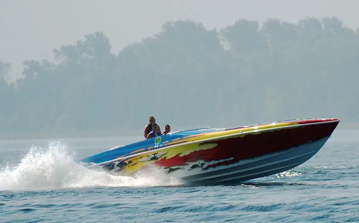 A colorful speedboat races across the water, throwing up a broad white wake, with three people aboard.