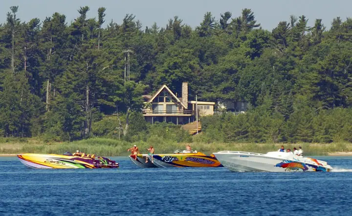 Three colorful speedboats travel side by side past a forested shoreline and a house.