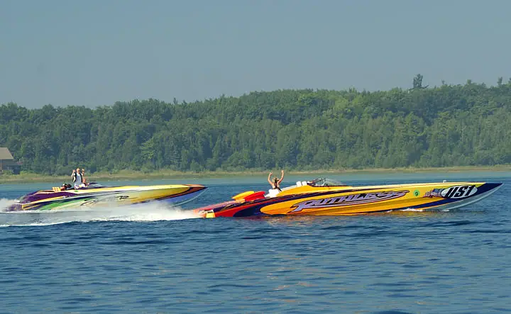 Two brightly colored speedboats race side by side, throwing wakes, with a forested shoreline behind them.