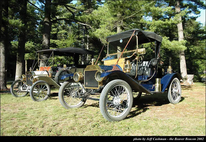 Two vintage touring automobiles with spoked wheels stand on a grassy clearing before tall pine trees.