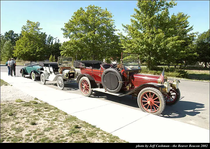 Three antique automobiles are displayed along a sidewalk beside a paved road, with people and trees in the background.