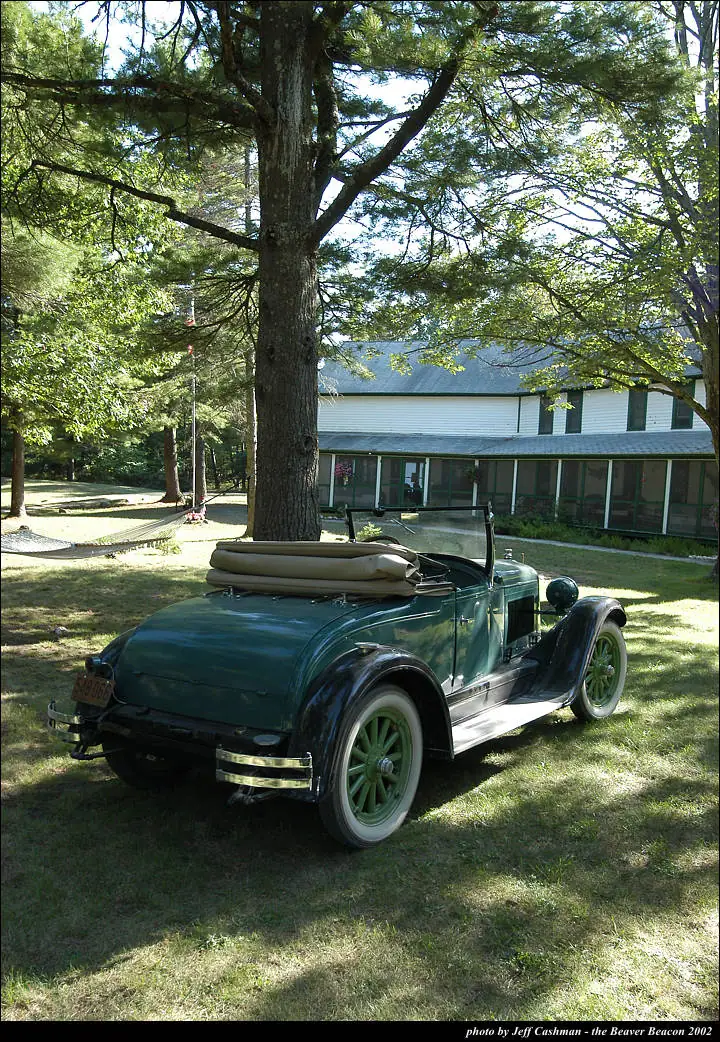 A green vintage convertible with its folded top rests beneath trees beside a long screened building.
