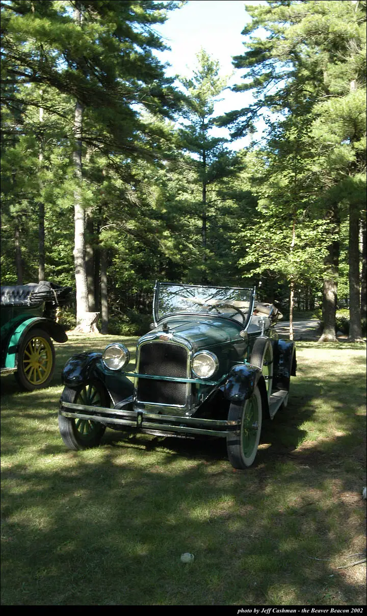 Dark green vintage convertible displayed on grass among tall pine trees, with another antique car partly visible.