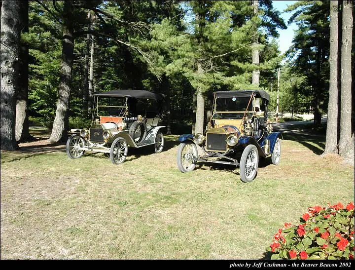 Two vintage automobiles are displayed on grass beneath pine trees, with red flowers in the foreground.