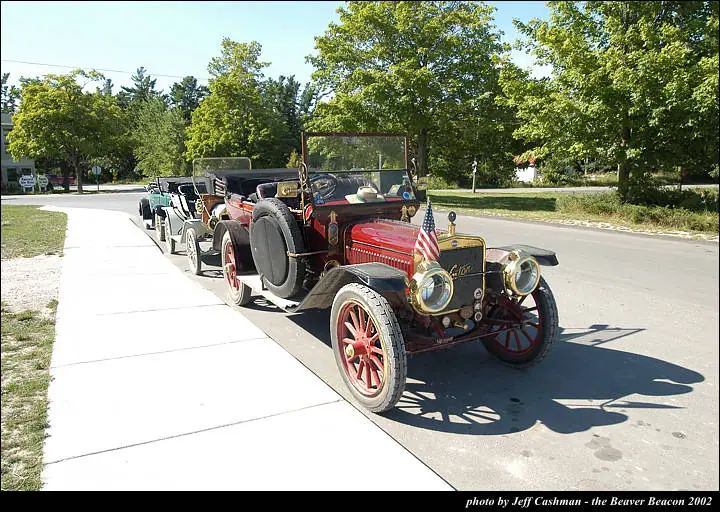A row of antique open automobiles is parked along a sidewalk, with a red car in front.
