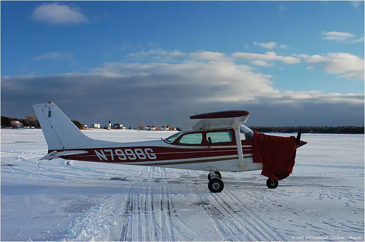 A small airplane with a red tarp over its nose rests on a snow-covered frozen surface.