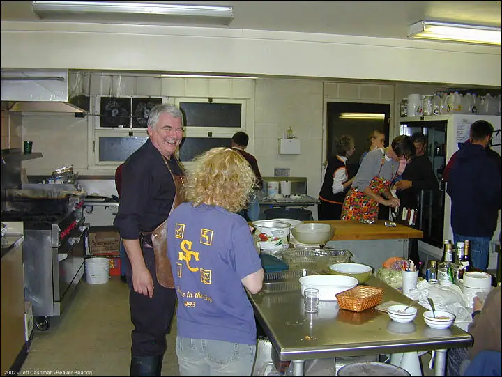 People prepare food in a busy kitchen around stainless-steel worktables filled with bowls and utensils.