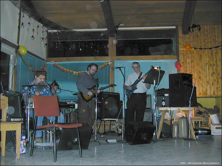 Three musicians perform with guitars and drums in a wood-paneled room decorated with spiders, balloons, and garlands.