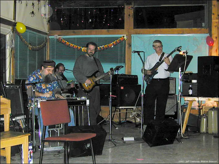 Four musicians perform indoors with a keyboard, guitars, microphones, and amplifiers.