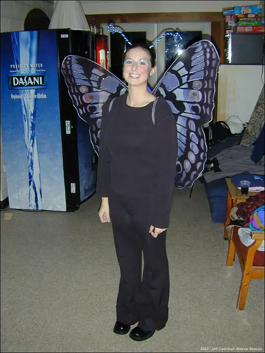 A smiling woman in a butterfly costume stands indoors beside a Dasani vending machine.