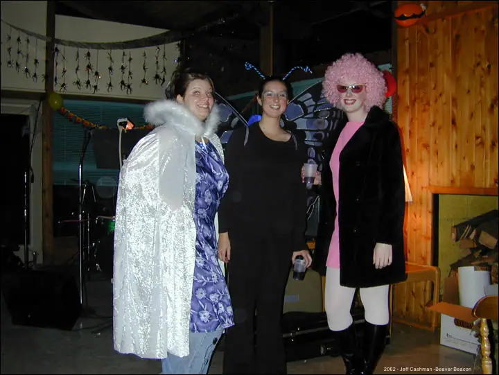 Three people in colorful costumes pose indoors beneath Halloween decorations.