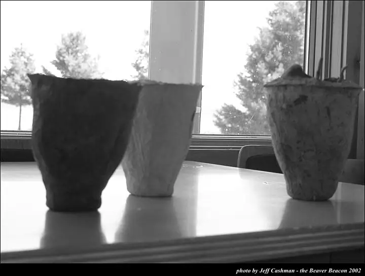 Three textured paper cup sculptures are displayed on a table before large windows.